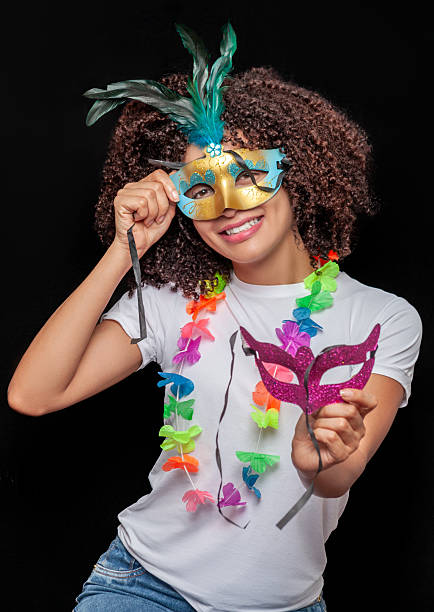 Vertical portrait of a woman smiling while holding a carnival mask decorated with feathers, wearing a colorful festive lei. Black background and studio lighting. Ideal for carnival, festivities,