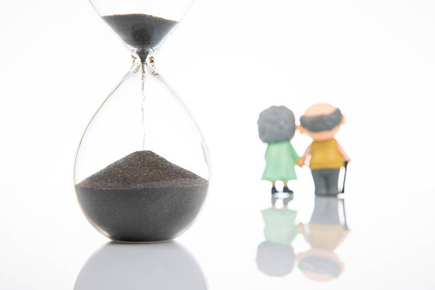 featuring two elderly figurines holding hands, gazing at a large hourglass with most sand in the top, symbolizing the passage of time and life’s fleeting nature. White background with reflective surface.