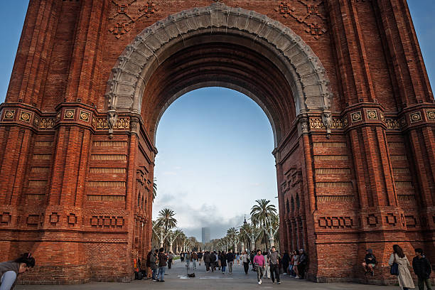 Barcelona, Spain - April 5, 2025: The photograph centers on Barcelona's Arc de Triomf, a red-brick triumphal arch that serves as a ceremonial gateway to the promenade on Passeig de Lluis Companys. Shot from beneath the arch, the image frames a palm-lined boulevard and an open sky, revealing the scale of the structure and the decorative Neo Mudejar details in stone and brick. Visitors stroll through the space, adding context and a sense of everyday city life. The scene highlights a celebrated Catalan landmark, urban public space, and historic architecture within Spain's most visited coastal metropolis.