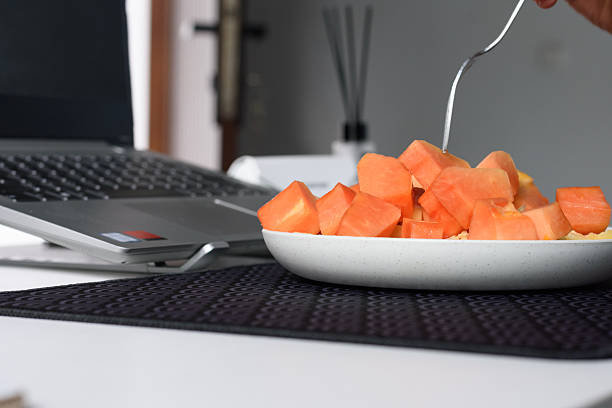 A close-up shot of person's hand uses a fork to pick up a piece of diced papaya from a white bowl. The bowl contains both orange papaya and yellow pineapple chunks and sits on a textured black mat on a white desk. The background is blurred, showing a laptop on a raised stand and other desk accessories. This image strongly emphasizes the action of taking a healthy work break, focusing on eating, productivity, and the integration of wellness into a modern work-from-home setting