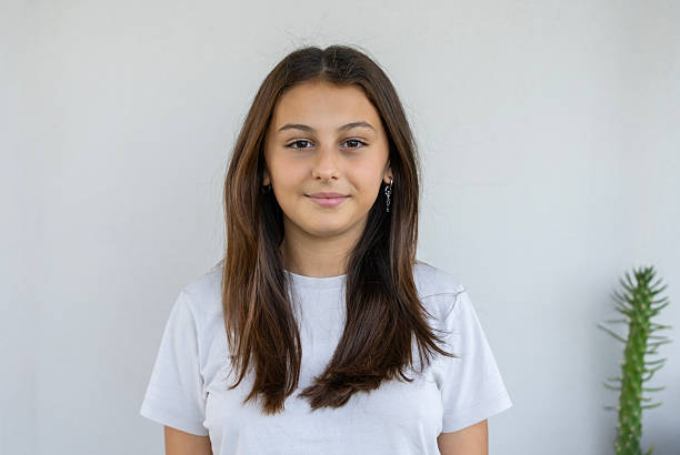 Portrait of a teen girl looking calmly at the camera while standing in front of a plain wall.