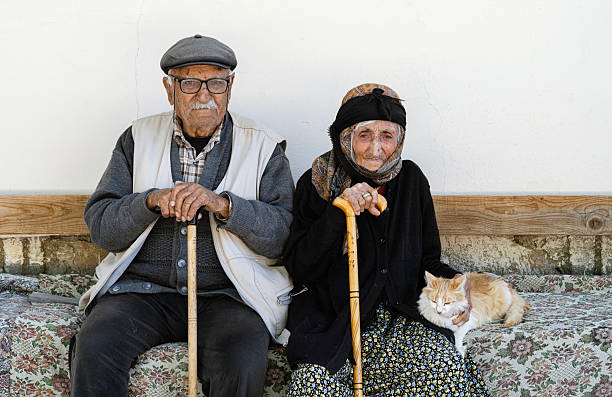 An elderly couple sitting side by side on a bench outside their rural home, each holding a wooden walking stick. The woman gently pets a cat resting on her lap