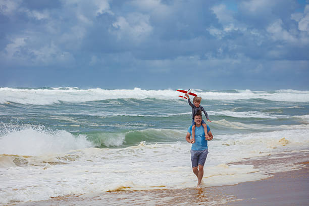 A man strolls along the shoreline with a child on his shoulders, holding a toy airplane under a partly cloudy sky