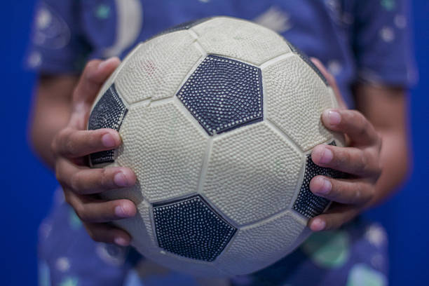 A focused child in a blue striped shirt prepares to kick a soccer ball during a sunny outdoor game near a goalpost.