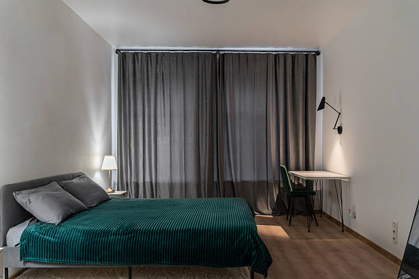 istock large bright bedroom with double bed. on bed is green bedspread and pillows. grey curtains on windows. table and chair near white wall. lamp on table. Photo of bedroom with different lighting. 2250082571