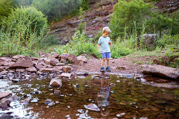 A cheerful toddler with blond hair stands by a clear mountain river, holding a small pebble while enjoying outdoor play. He is dressed in a T-shirt and shorts, surrounded by greenery and rocky terrain, capturing a peaceful moment of childhood exploration in nature.