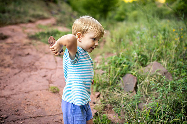 Side view of a little blonde boy, around two to three years old, standing on a mountain trail and tossing small pebbles. Wearing a T-shirt and shorts, he is fully immersed in his play. The scene is surrounded by lush green nature, capturing a peaceful and carefree moment of childhood exploration.