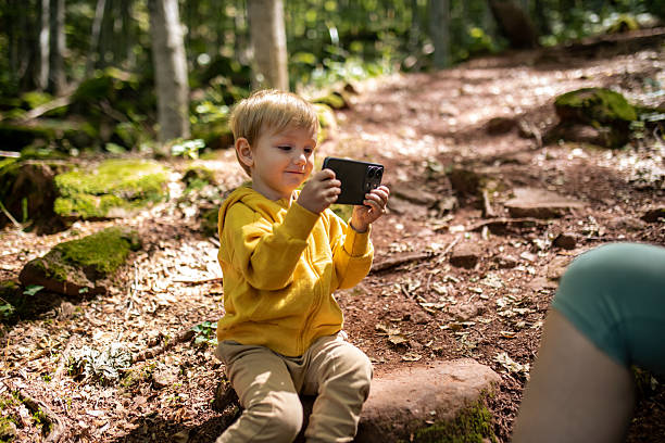 A blond toddler is playfully holding a smartphone, trying to take a photo by himself on a mountain forest trail. He is dressed in relaxed hiking clothing, surrounded by lush green nature and sunlight filtering through the trees. The scene captures a moment of curiosity, playful exploration, and independent learning in the outdoors.