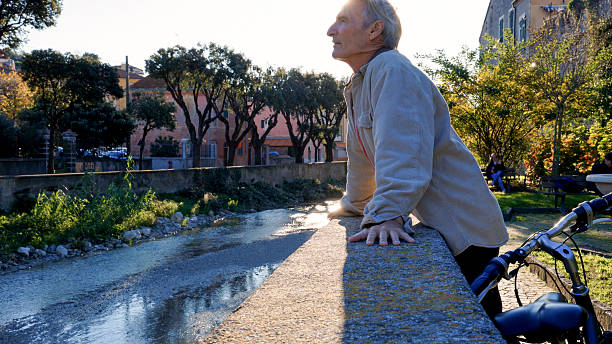 Senior man leans against wall above river, bicycle beside him, Finale Ligure