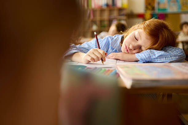 Redhead schoolgirl feeling exhausted while writing in a textbook during a class at elementary school.