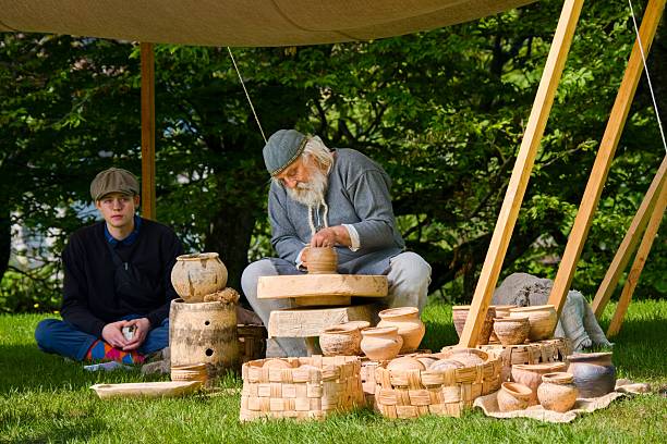 Grobina, Latvia, May 31, 2025 - Traditional potter shaping clay vessels outdoors during a historical craft demonstration, surrounded by handmade pottery and natural greenery.