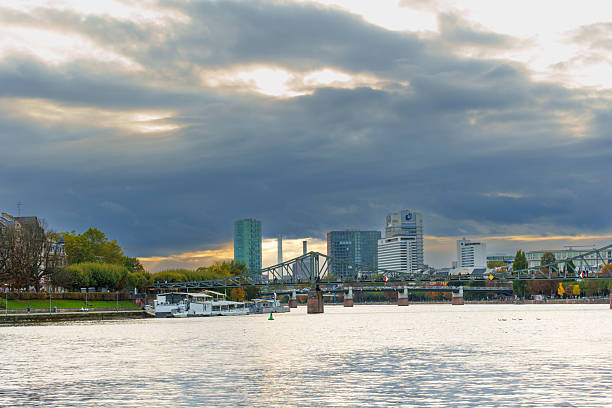 Frankfurt, Germany - October 21, 2025: Cloudy sky with rays of sunlight illuminate Frankfurt's iconic skyline, bridge, and the serene Main River, creating a picturesque autumn cityscape scene.