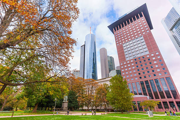 Frankfurt, Germany - October 21, 2025: Vibrant autumn foliage in an urban park contrasting Frankfurt's impressive skyline view, featuring prominent modern skyscrapers under a bright, cloudy sky.