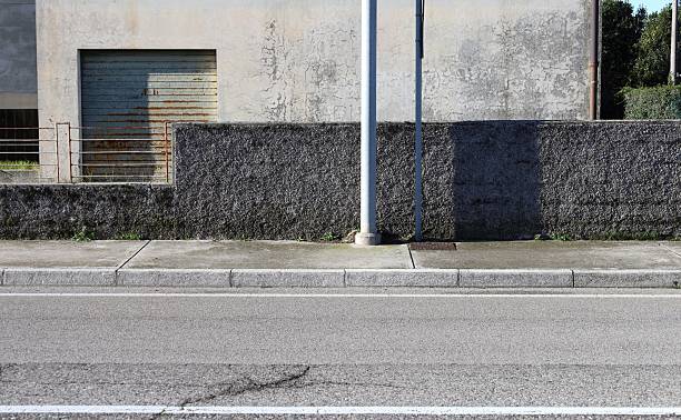 istock Pole on sidewalk with rough concrete fence and rusty railing. Abandoned building on behind, street in front 2250099666