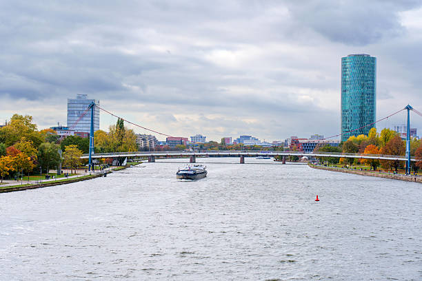 Frankfurt, Germany - October 21, 2025: Urban panorama capturing Frankfurt's Westhafen Tower and Holbeinsteg bridge over Main River. A cargo vessel traverses the waterway amidst autumn foliage.