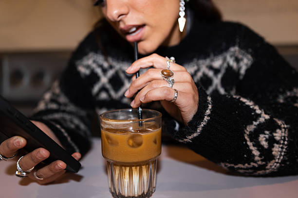 A close-up view of a beautiful young woman sitting in a bar in Newcastle Upon Tyne, England. She is scrolling on her smartphone while taking a sip of her iced coffee. She is wearing a knitted jumper.