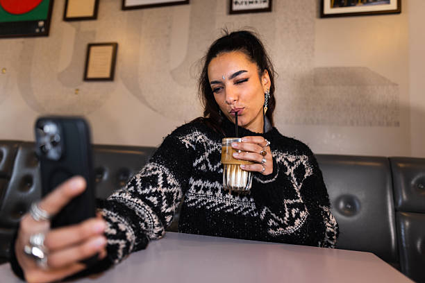 A medium view of a beautiful young woman sitting in a bar in Newcastle Upon Tyne, England. She is taking a selfie on a smartphone while taking a sip of her iced coffee. She is wearing a knitted jumper.