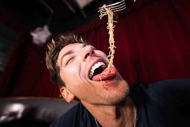 A medium view of a caucasian man sitting at a table at a restaurant in Newcastle Upon Tyne, England. He is enjoying a selection of delicious foods and drinks. He is wearing fashionable clothing and smiling while having a great time. He is holding a fork up in the air, feeding noodles into his mouth.