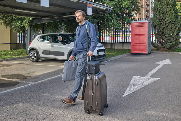A man walks through an outdoor parking lot carrying travel luggage, a backpack, and a small bag, preparing for a trip.