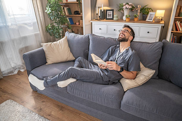 istock Healthcare worker relaxing on sofa reading book and laughing 2250120997