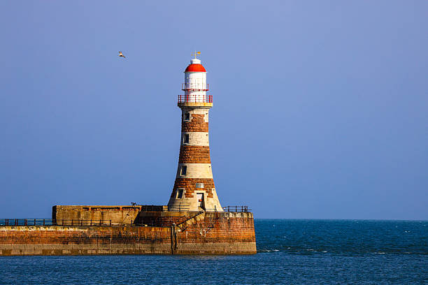 A striking stone lighthouse stands along a breakwater, illuminated by soft sunlight against a clear blue sky. Surrounded by calm ocean water, this peaceful coastal scene captures the timeless beauty and purpose of maritime architecture. Ideal for travel, nautical, and landscape themes.