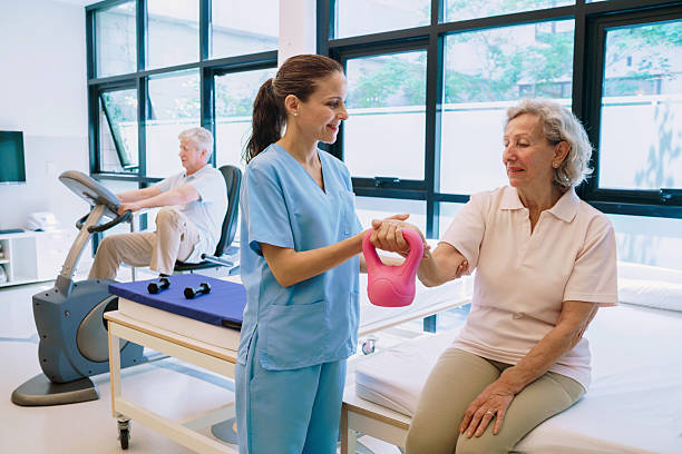 A friendly physiotherapist guides an elderly woman through a hand-guided exercise with a pink kettlebell in a rehabilitation setting. Background shows a man using a stationary bike, highlighting care, fitness, and recovery.