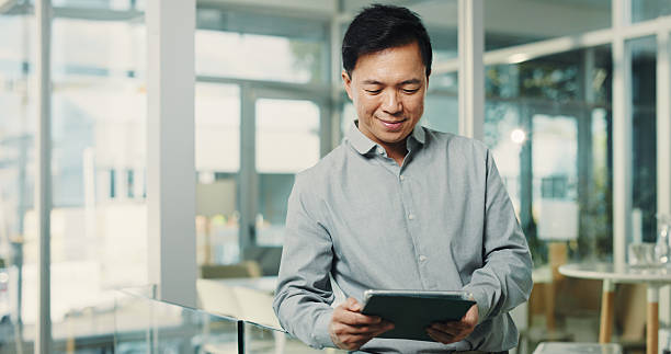 istock Asian man, reading and tablet in office for research, editing article and schedule update of draft. Person, digital and smile for breaking news, proofreading story and interview notes for publication 2250132264