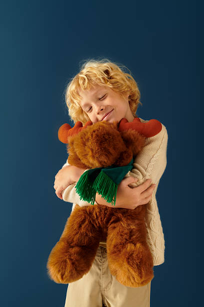 Blonde boy with wavy hair joyfully hugs a plush moose in a studio setting