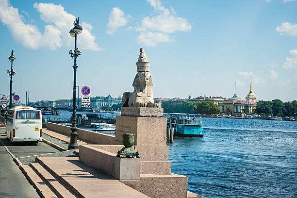 Ancient Egyptian sphinx statue in Saint Petersburg city with the Neva river, the Admiralty building, a tour boat, and a bus in the background on a sunny day