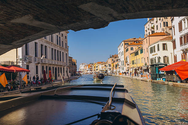 Venice, Italy - October 8, 2025: Boat moving under a bridge, navigating a vibrant canal lined with historic buildings and people along the waterfront in Venice, Italy