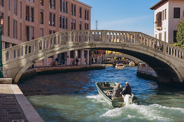 Venice, Italy - October 10, 2025: Elderly couple with a dog enjoying a boat ride along a canal, passing under a historic arch bridge in Venice, Italy