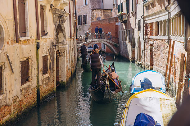 Venice, Italy - October 10, 2025: Gondoliers guiding tourist gondolas along a narrow Venetian canal, passing under a bridge and historical buildings