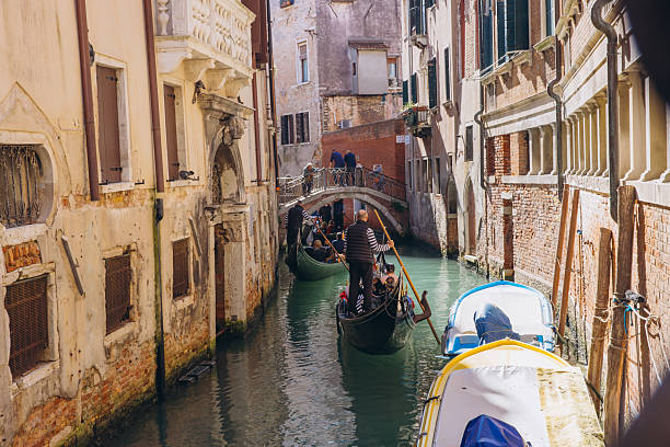 Venice, Italy - October 10, 2025: Gondoliers punting tourists navigate a narrow canal in Venice, Italy, framed by historic buildings and a small bridge
