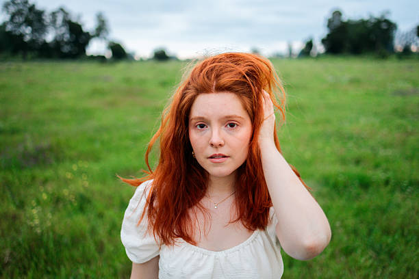 A close portrait of a young red-haired woman standing in a green meadow, gently holding her hair while looking forward. The natural gesture and soft outdoor light convey introspection, vulnerability, and emotional presence