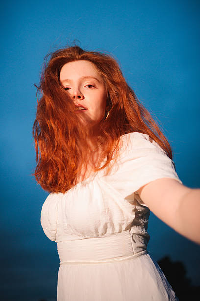A low-angle portrait of a red-haired woman in a white dress reaching toward the camera beneath a deep blue evening sky. The dynamic perspective and dramatic lighting suggest connection, vulnerability, and expressive movement
