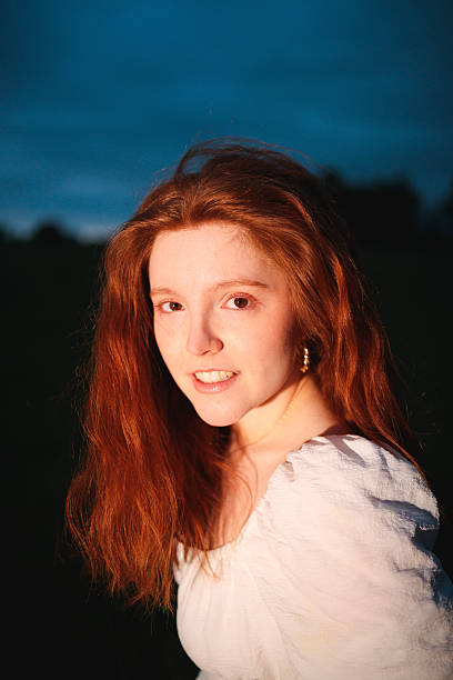 A close portrait of a red-haired woman in a white dress gazing toward the camera as warm light contrasts with the deep blue evening sky. Her composed expression suggests presence, confidence, and emotional balance