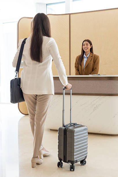 Hotel guest receives an access card from a receptionist while checking in at the front desk. A clear protective barrier separates them, ensuring safety and professionalism. In the background, another guest waits her turn, and the modern, warmly lit hotel lobby creates a welcoming atmosphere for travellers arriving at their destination.