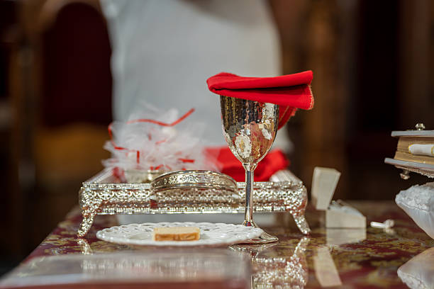 A silver cup sits on a decorative tray with a red napkin on top. Nearby, a plate holds a small pastry. Decor around suggests a formal gathering in a large hall.