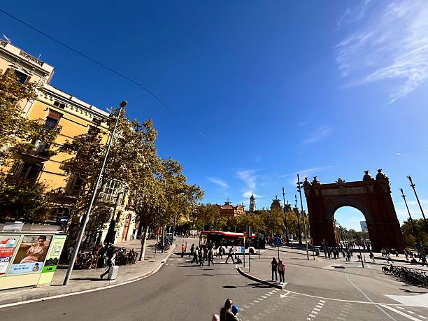 Barcelona, Cataluña, Spain - October 24, 2025: Arc de Triomf in Barcelona framed by blue summer sky, people crossing a busy intersection and strolling the promenade near Passeig de Lluís Companys landmark