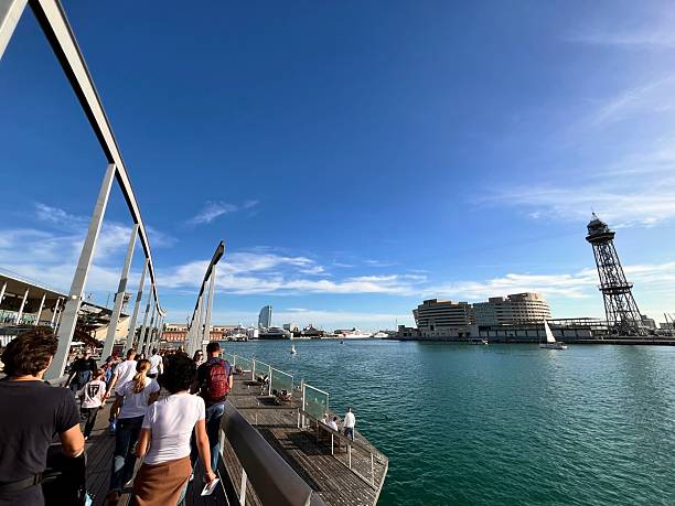 Barcelona, Cataluña, Spain - October 24, 2025: People are walking on the wooden bridge of Rambla de Mar, enjoying a sunny day with views of Port Vell, the cable car tower, and W Barcelona hotel in the distance