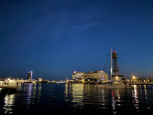 Barcelona, Cataluña, Spain - October 26, 2025: Waterfront view of Barcelona's Port Vell at twilight, featuring the illuminated cable car tower  and a catamaran cruising the calm waters with city lights reflecting