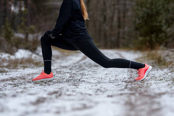 istock Focused on physical activity: Side view of a female runner performing a deep lunge to stretch the hamstring and hip flexor muscles on a snowy forest path. 2250178971
