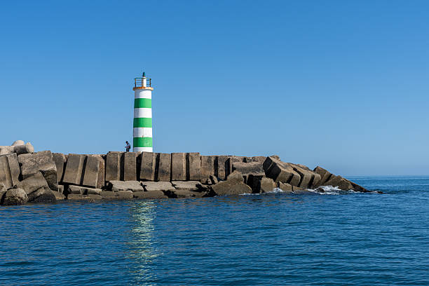 A lighthouse stands tall on a stone pier, guiding ships safely to shore on a sunny day