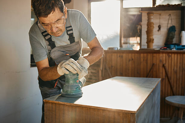 Mature man wearing overalls and safety glasses sanding wood in a sunny workshop
