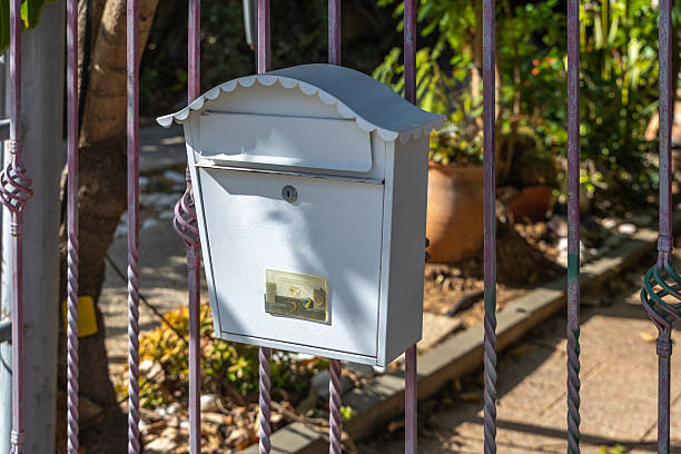 Close view of a white metal mailbox mounted on an iron fence at a residential home with garden foliage in sunlight, symbol of mail delivery and neighborhood communication
