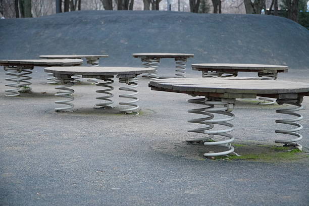 Row of vintage spring-mounted circular bouncers on concrete playground surface with large metal coil springs under wooden seats designed for children jumping play in outdoor public park. High quality photo
