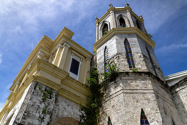 A detailed upward view of a historic colonial-era church tower in Bohol, Philippines. The stone walls, stained glass windows, and architectural details reflect Spanish influence, while lush green plants grow along the weathered facade. Captured under a bright blue sky, this image is ideal for themes of history, architecture, heritage landmarks, tourism, and Southeast Asian culture.