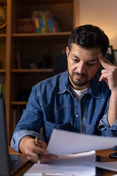 Focused man calculating finances at home, managing budget and reviewing important documents, experiencing stress and worry while working late from remote office