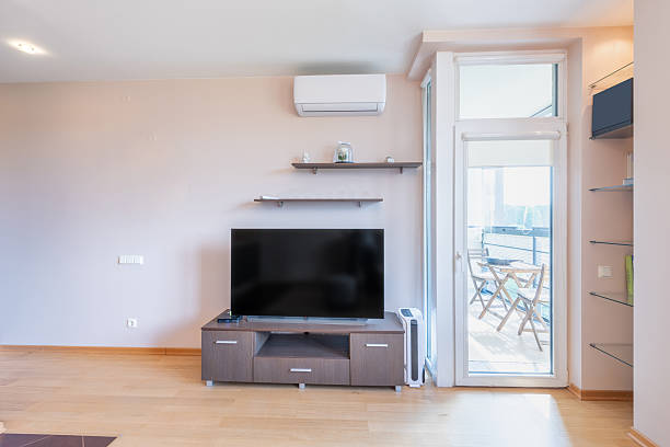 A large living room with a black television and a clock on the wall. The room is filled with white furniture and has a modern feel