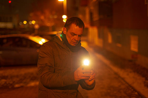 Man holding torch and phone on dimly lit urban sidewalk at night, wearing heavy coat, focused gaze, warm flashlight glow illuminating face, parked cars and blurred amber streetlights in background,